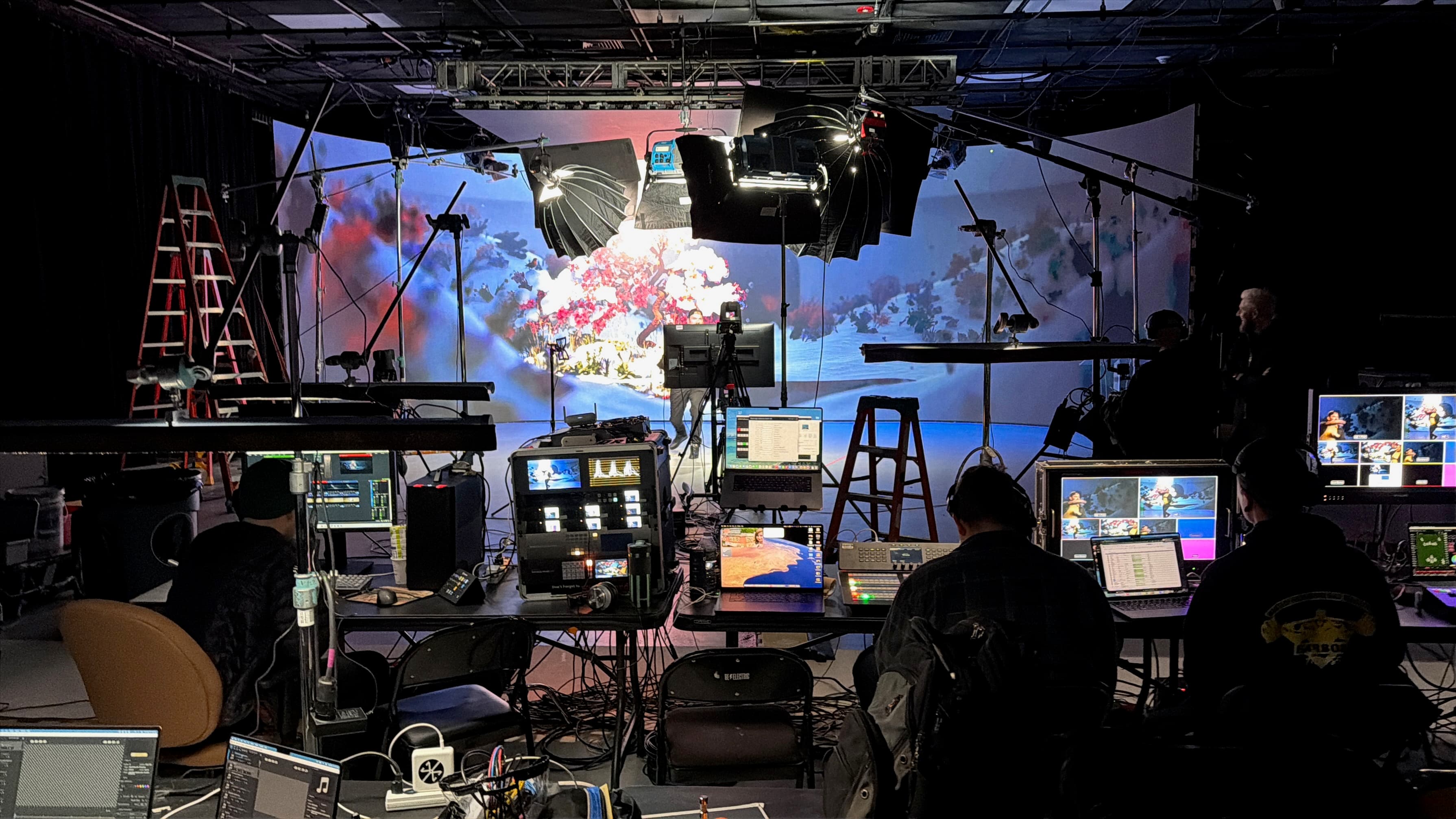 A live production in progress in a darkened studio: crew in headsets at a bank of tables running a vision mixer, laptops and a multiviewer of camera feeds, facing a large curved video wall showing an underwater coral scene, with lighting panels and truss rigged overhead.