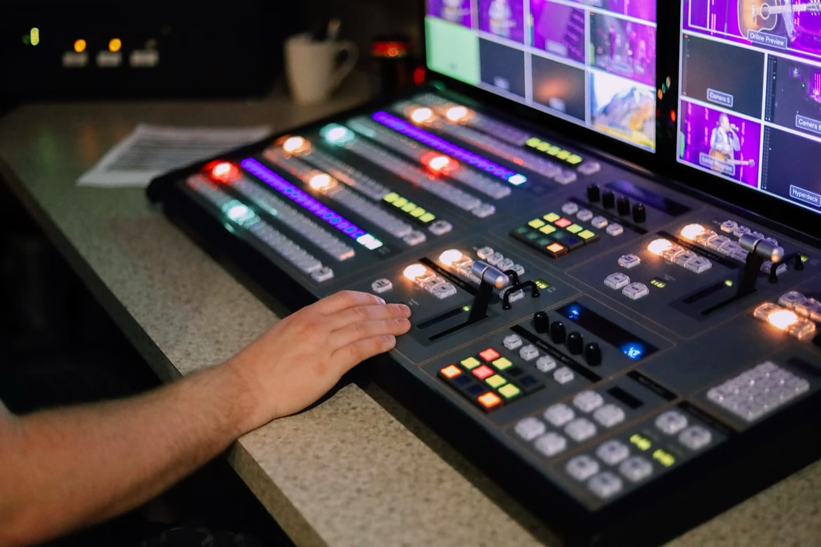 An operator's hand resting on a lit vision-mixer panel, with a multiviewer above it showing camera feeds from a concert