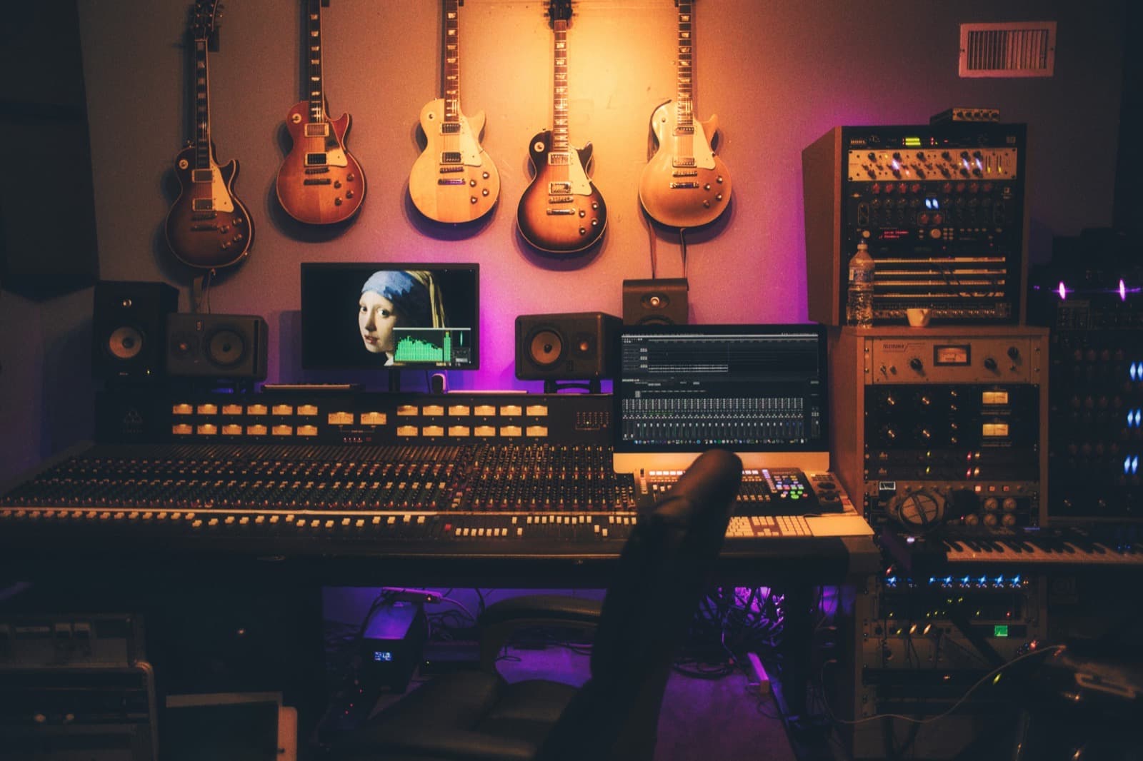 A large mixing console in a studio, with outboard racks either side and guitars hung on the wall behind